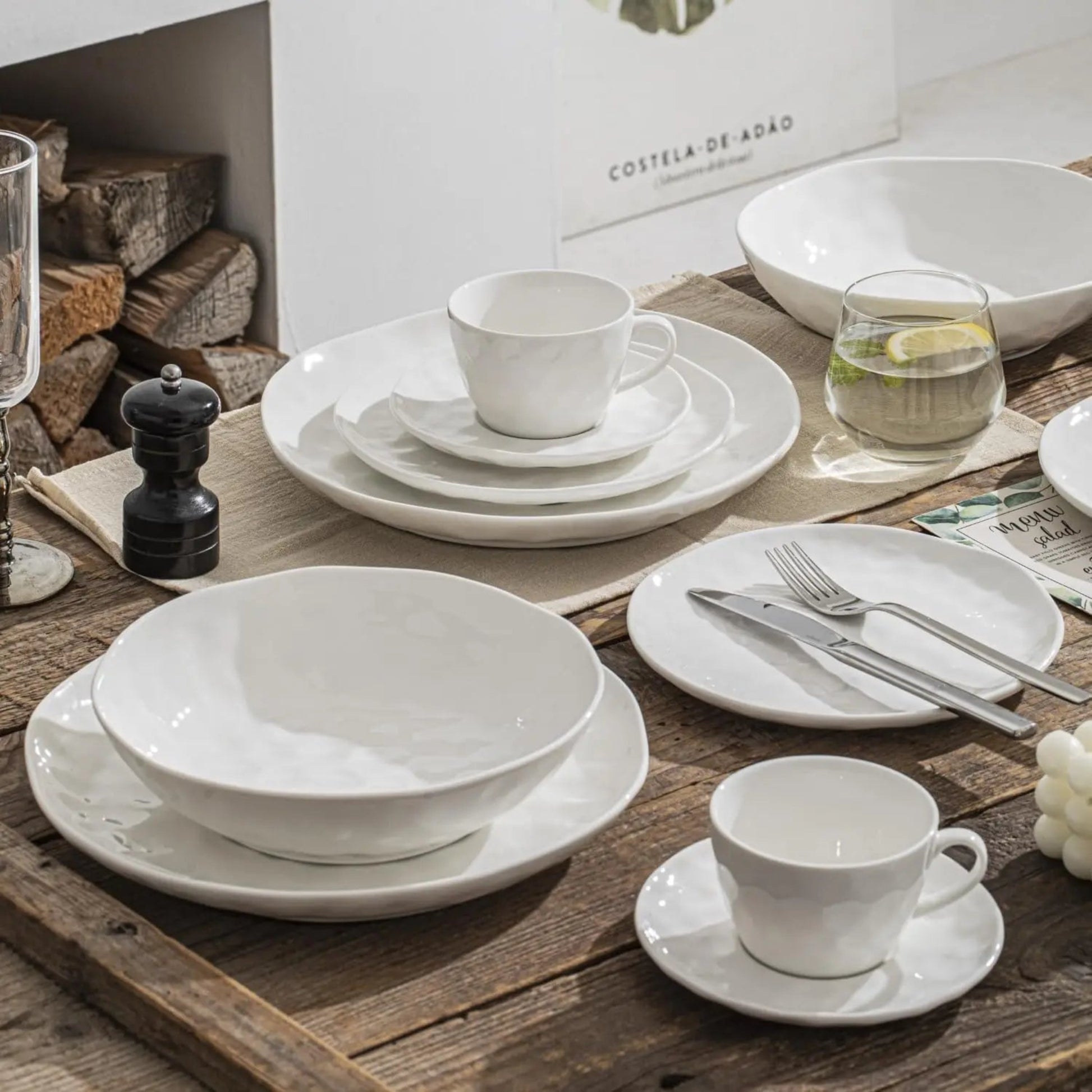 White ceramic dinnerware set on a wooden table with a 'Costela de Adão' branded box in the background.