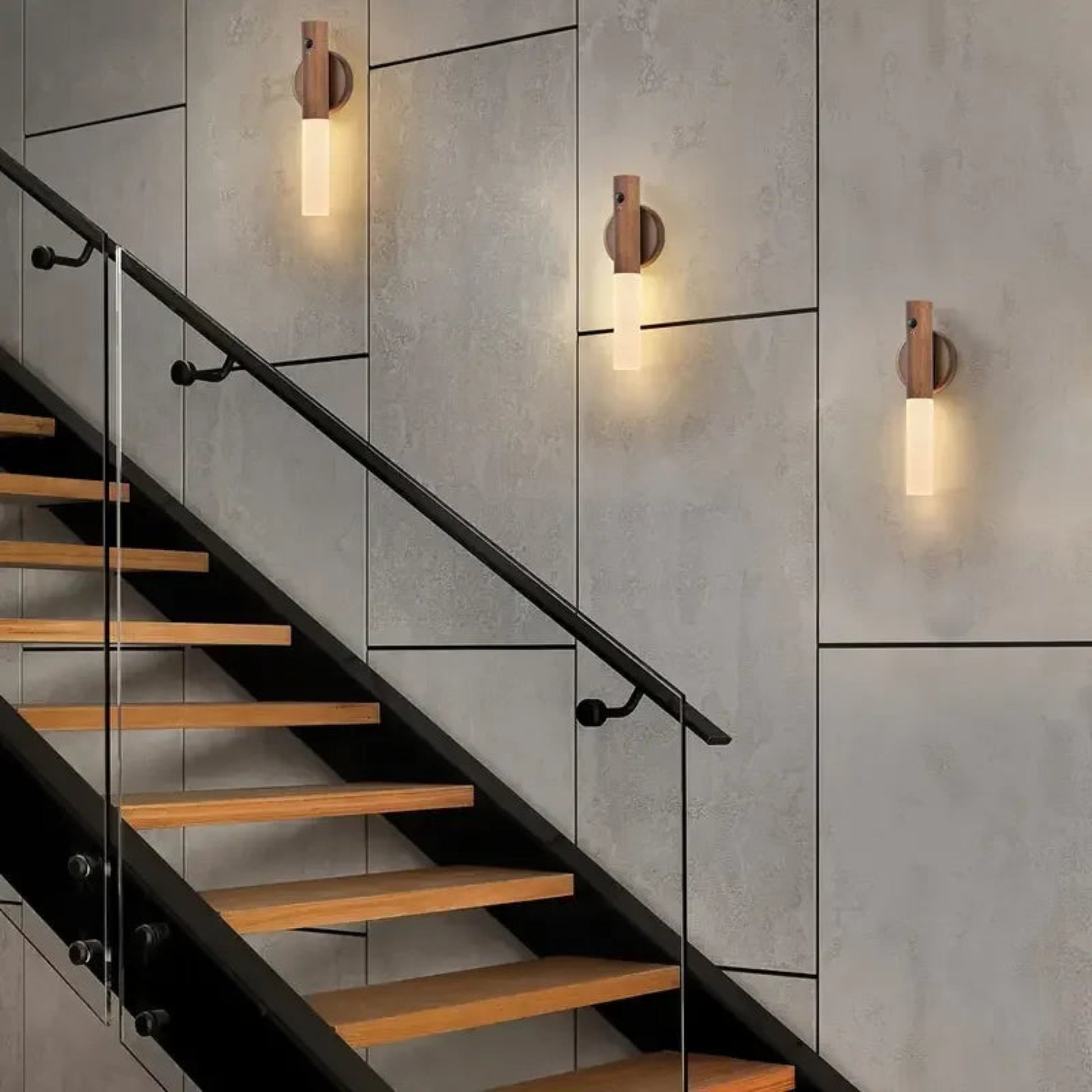 Modern staircase with wooden steps and glass railings against a concrete wall with wall lights.