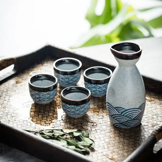Set of four ceramic cups and a bottle on a woven mat with greenery in the background