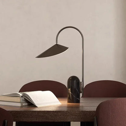 Modern table lamp on a wooden table with an open book and decorative bottle against a plain wall.