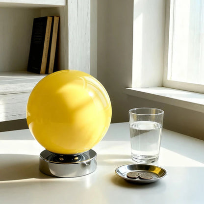 Yellow glass orb on a stand next to a glass of water and coins on a light surface with a window in the background.