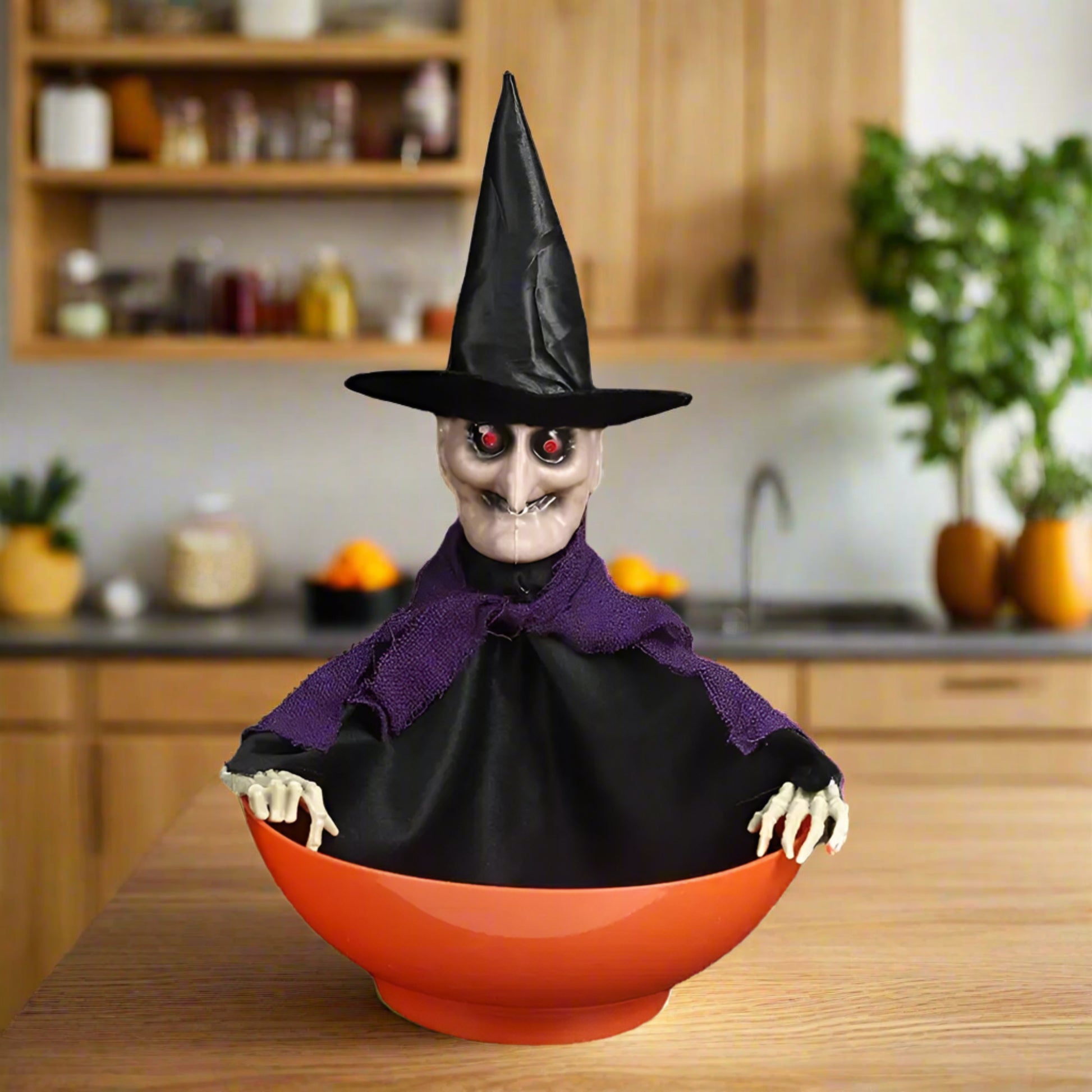 Halloween decoration of a witch in a black hat and purple cape with red eyes, sitting inside an orange bowl.