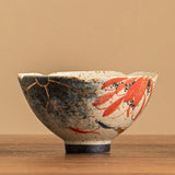 A handmade ceramic bowl with a scalloped rim, featuring a mottled blue and white glaze with gold leaf crackle repair patterns and red floral motifs, set on a wooden surface against a warm beige background.