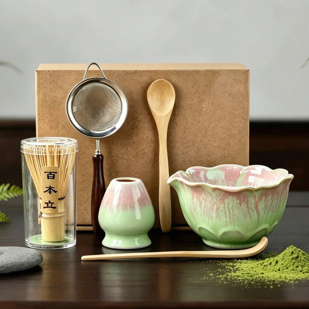 A Japanese matcha tea preparation set displayed on a dark wooden table in front of a brown kraft box. The set includes a pink and green ceramic matcha bowl, a matching small vase, a bamboo whisk in a clear glass container, a wooden spoon, a metal sieve, and a bamboo scoop resting on the bowl with green matcha powder scattered in front.