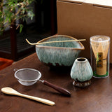 Tea-making set with ceramic bowl, whisk, and other tools on a wooden surface.