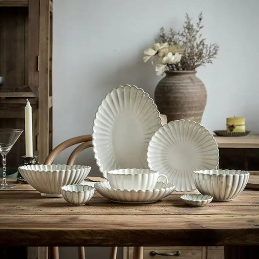 Set of white ceramic dishes on a wooden table with a vase and candle in the background.