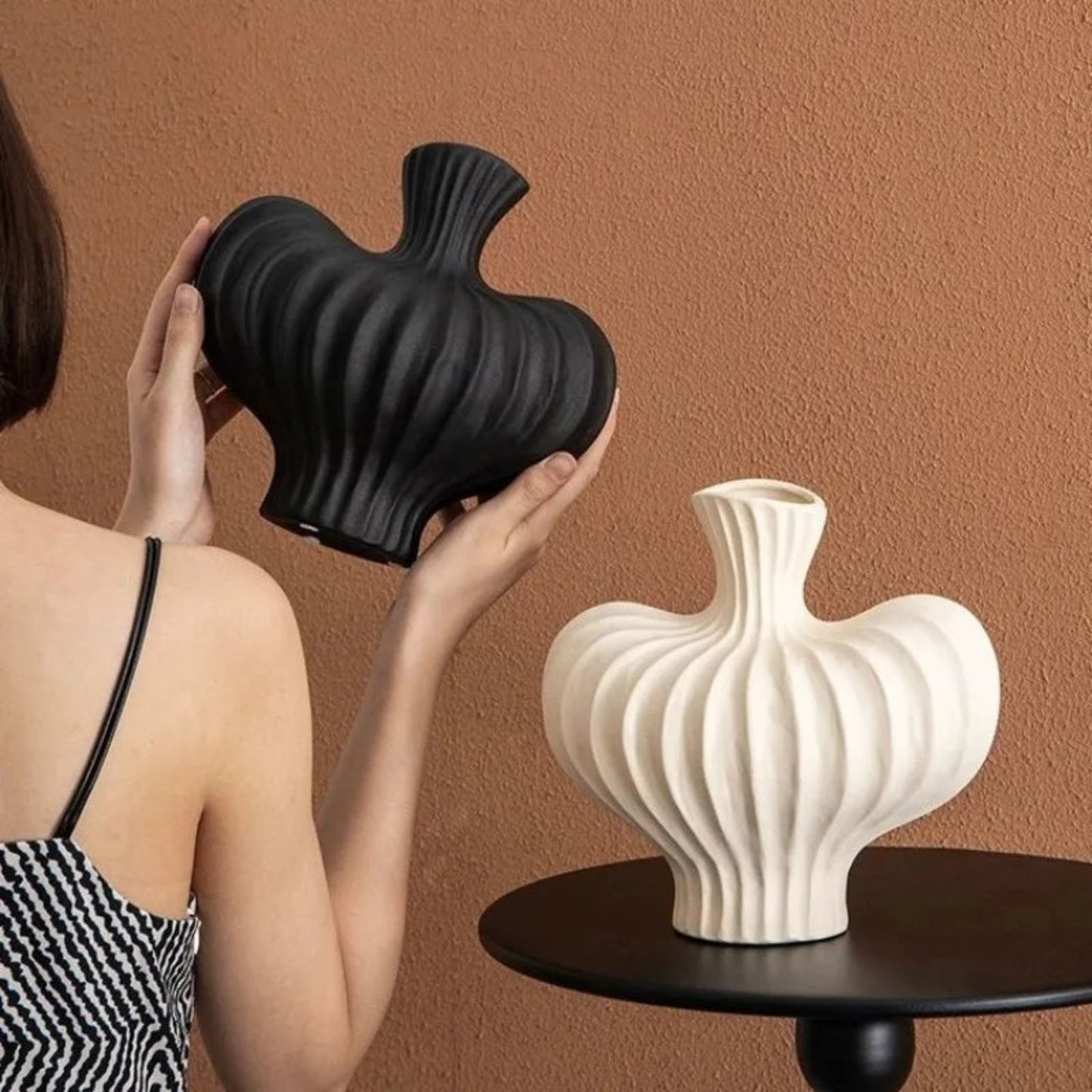 Person holding a black textured vase next to a white vase on a table against a brown wall.