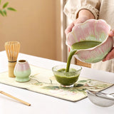 Person pouring green liquid from a pink and green bowl into a glass on a table with tea-making tools.
