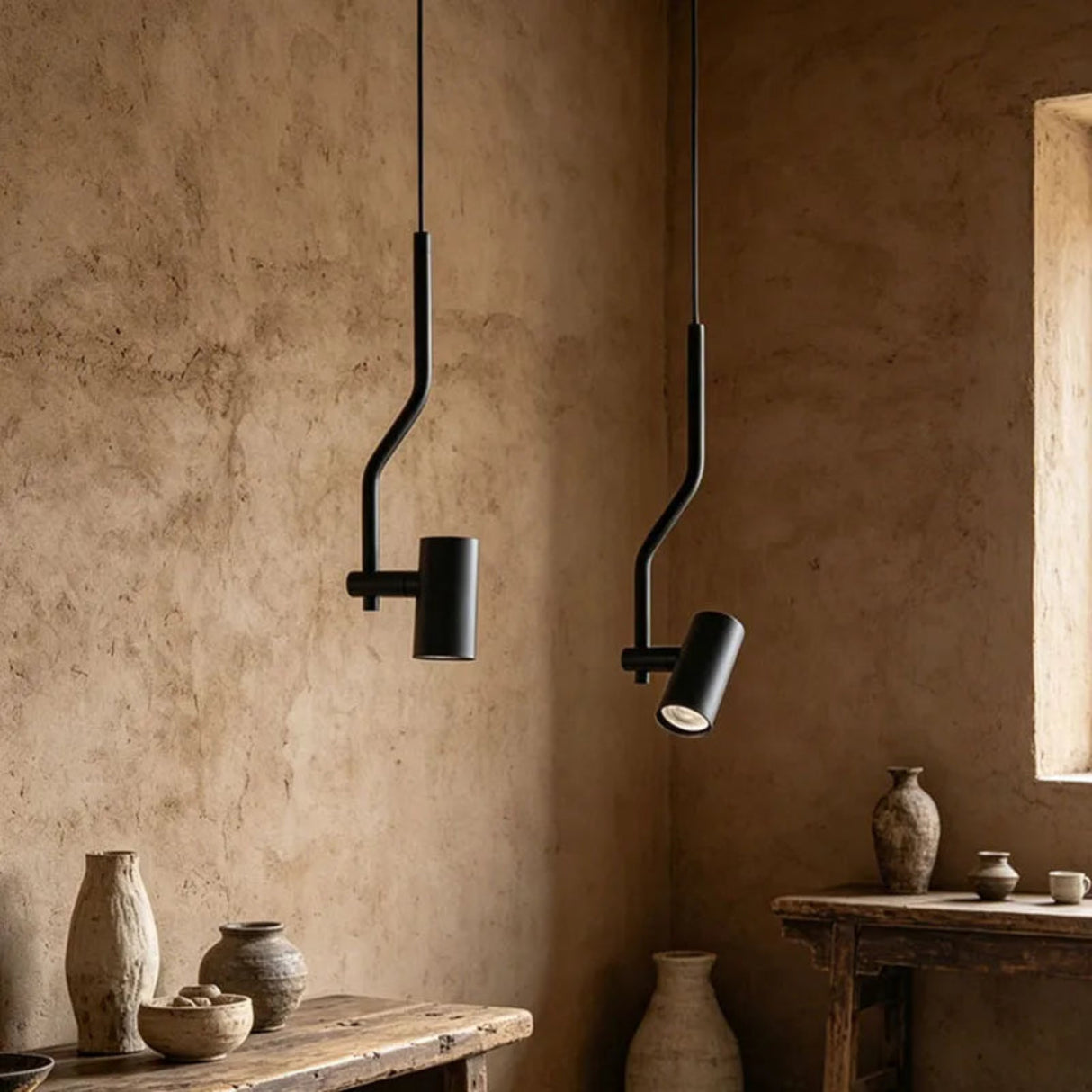 Two modern black pendant lights with curved, bent metal arms hang in a rustic room with textured beige plaster walls and wooden tables holding ceramic vases.