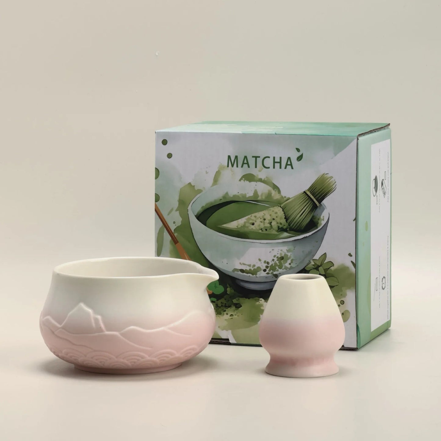 Matcha tea set with a pink bowl and whisk in front of a matcha-themed box on a beige background