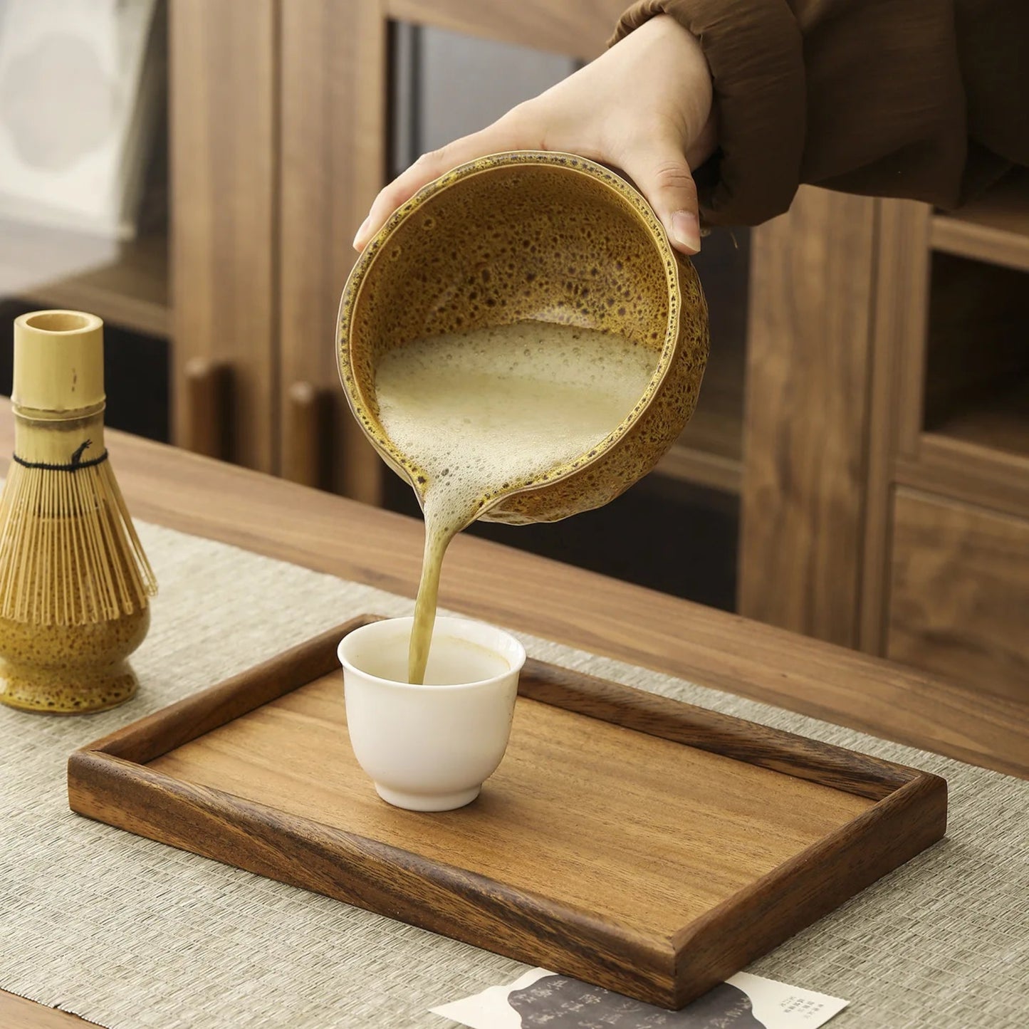 Person pouring a liquid from a textured bowl into a white cup on a wooden tray.