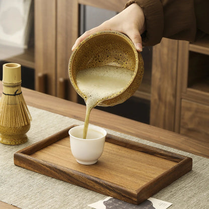 Person pouring a liquid from a textured bowl into a white cup on a wooden tray.