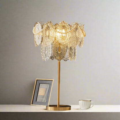 Decorative table lamp with intricate design on a stand, placed on a surface with a dark wall background.
