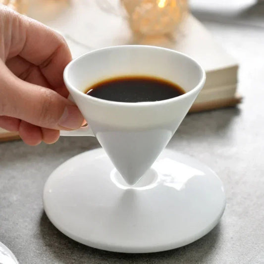 White ceramic coffee cup with a handle, filled with dark liquid, held by a hand on a light surface.