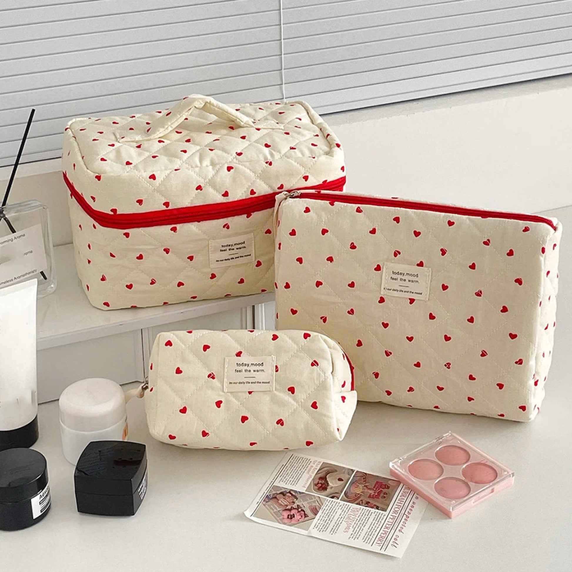 Set of three beige cosmetic bags with red heart patterns on a white surface.