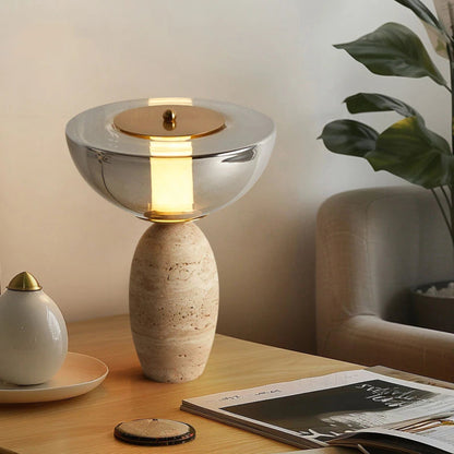 Table lamp with a ceramic base and glass shade on a wooden surface, with a plant and magazine in the background.