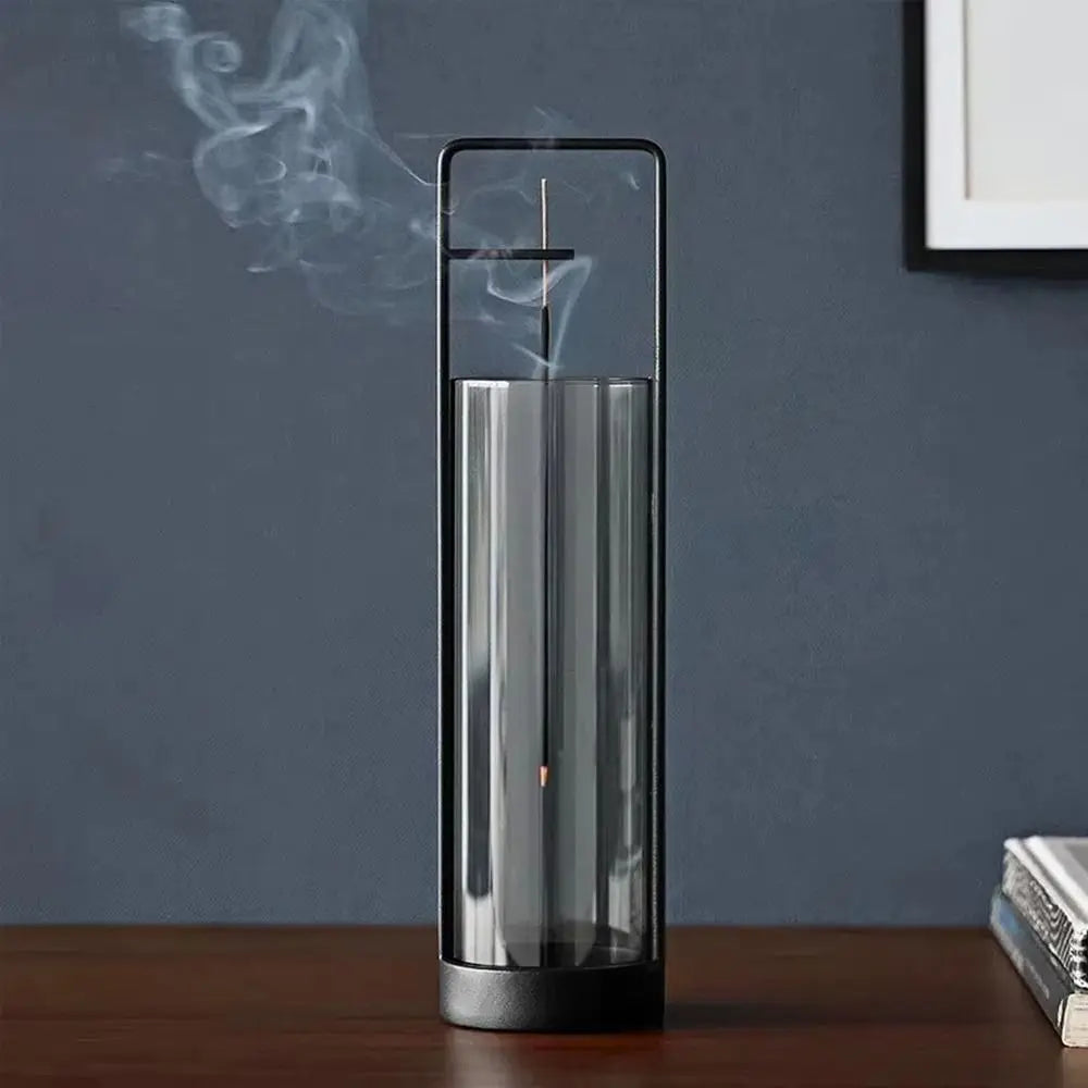 A modern cylindrical incense burner with a clear glass body, black metal frame, and black base, shown on a wooden table with incense smoke rising against a dark blue-gray wall.
