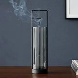 A modern cylindrical incense burner with a clear glass body, black metal frame, and black base, shown on a wooden table with incense smoke rising against a dark blue-gray wall.