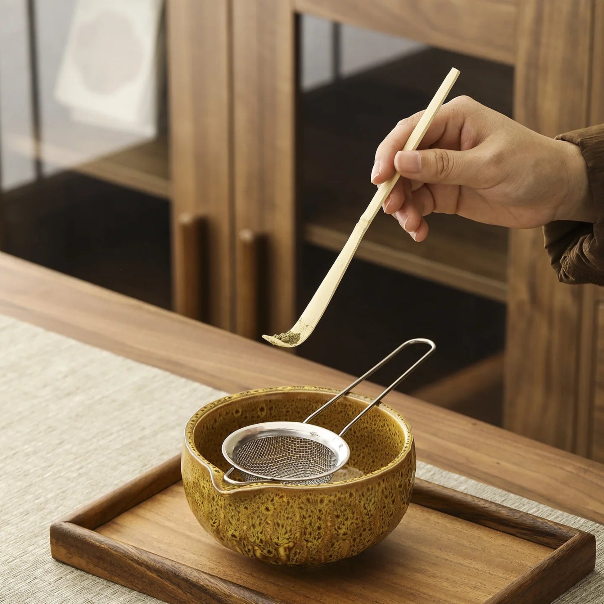 Person using a wooden spoon to stir tea in a ceramic bowl on a wooden tray.