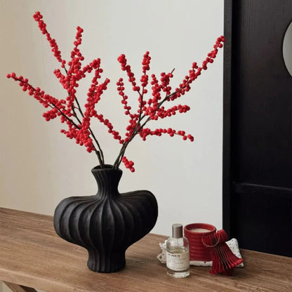Black vase with red branches on a wooden surface next to a candle and small bottle.