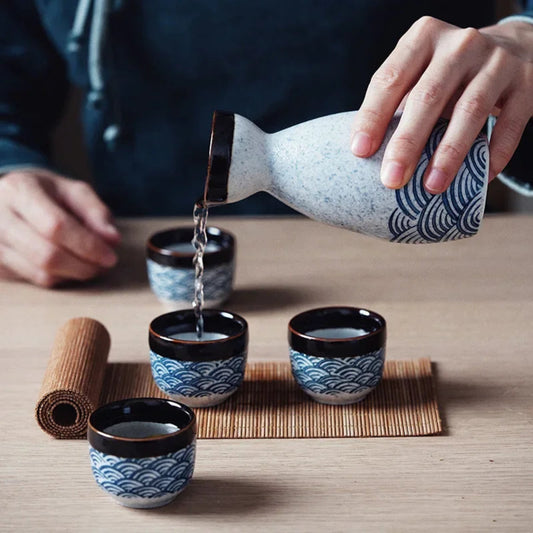 Person pouring sake from a traditional Japanese sake bottle into four small cups on a wooden table.