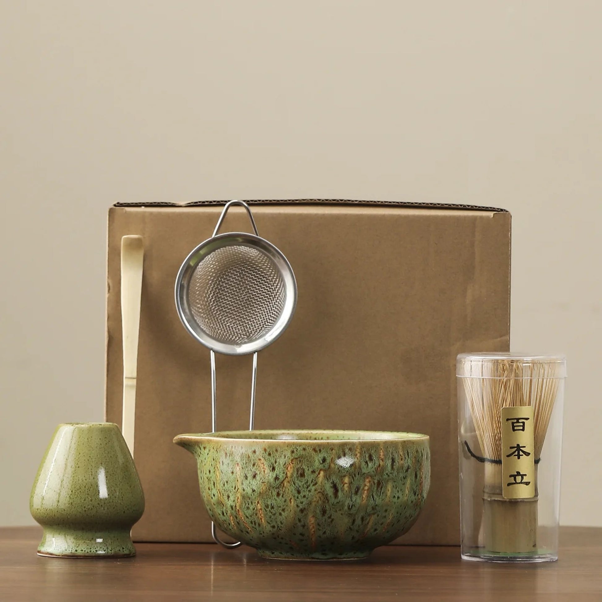 Tea-making set with green ceramic bowl, strainer, whisk, and glass on a beige background