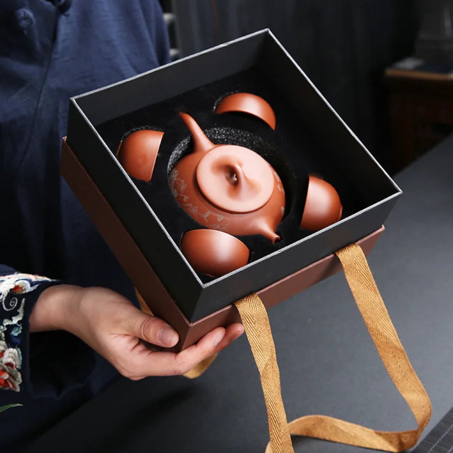 Person holding a black gift box with a ceramic teapot and cups inside.