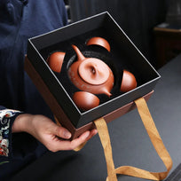 Person holding a black gift box with a ceramic teapot and cups inside.