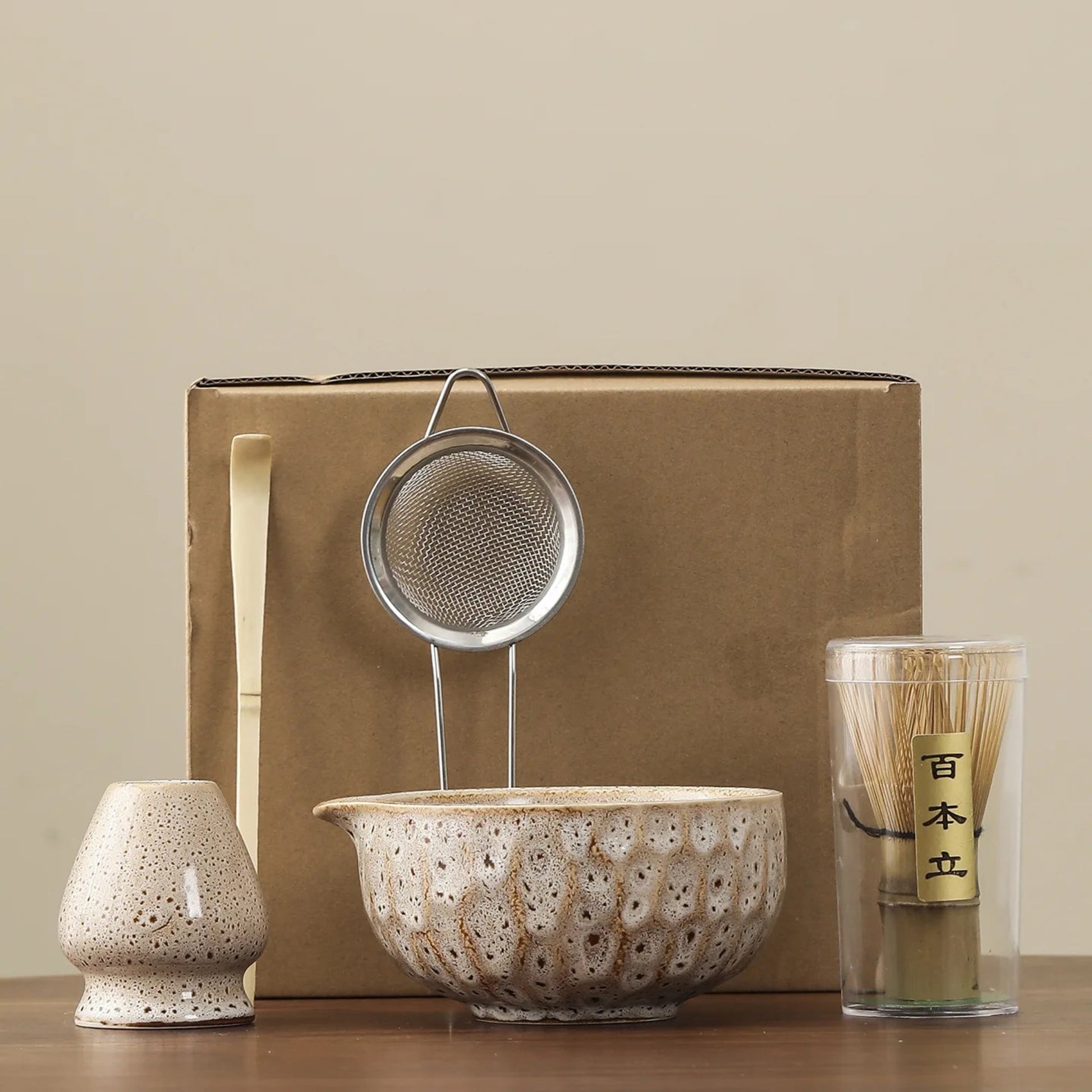 Set of ceramic and metal items including a bowl, strainer, and container with a wooden stick on a beige background.