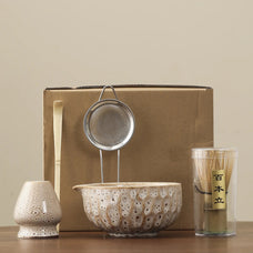 Set of ceramic and metal items including a bowl, strainer, and container with a wooden stick on a beige background.
