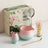 Matcha tea set with pink and green bowl, small cup, whisk, and box on a white surface.