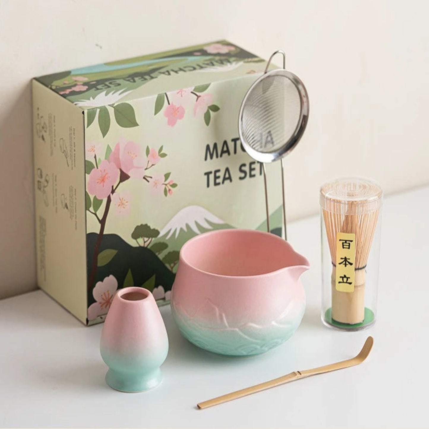 Matcha tea set with pink and green bowl, small cup, whisk, and box on a white surface.