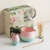 Matcha tea set with pink and green bowl, small cup, whisk, and box on a white surface.
