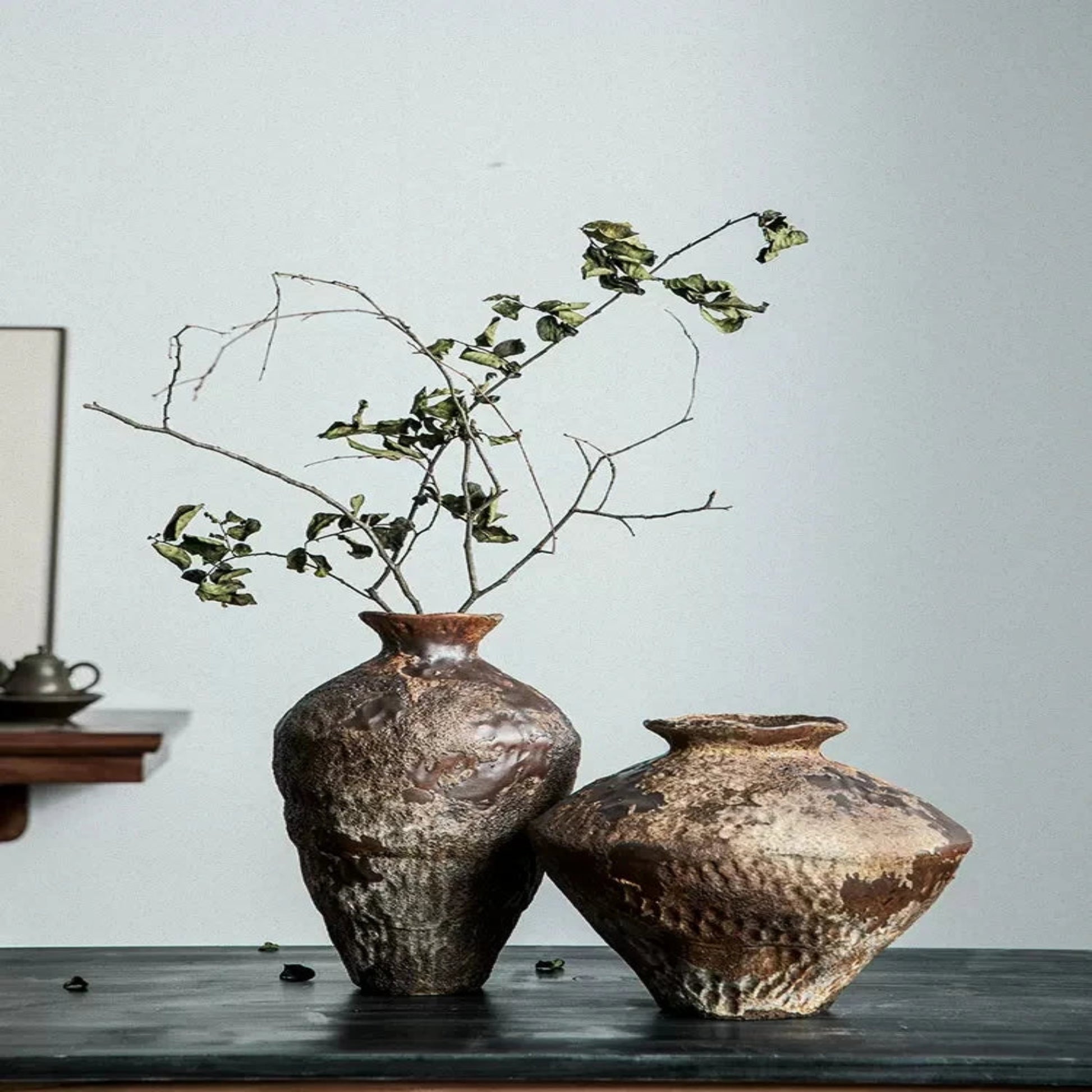 Two rustic ceramic vases with dried branches on a wooden surface against a light gray wall.