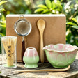 Tea-making set with a green ceramic bowl, wooden spoon, and other tools on a wooden surface with a natural background.