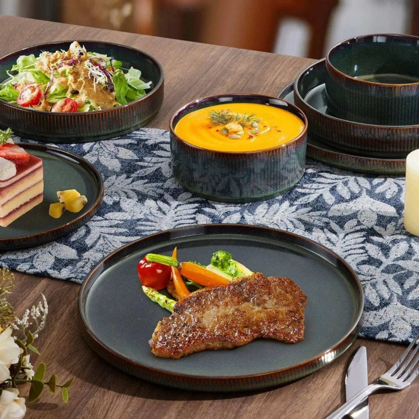 Dinner table setting with a steak, soup, salad, and bread on a patterned tablecloth.
