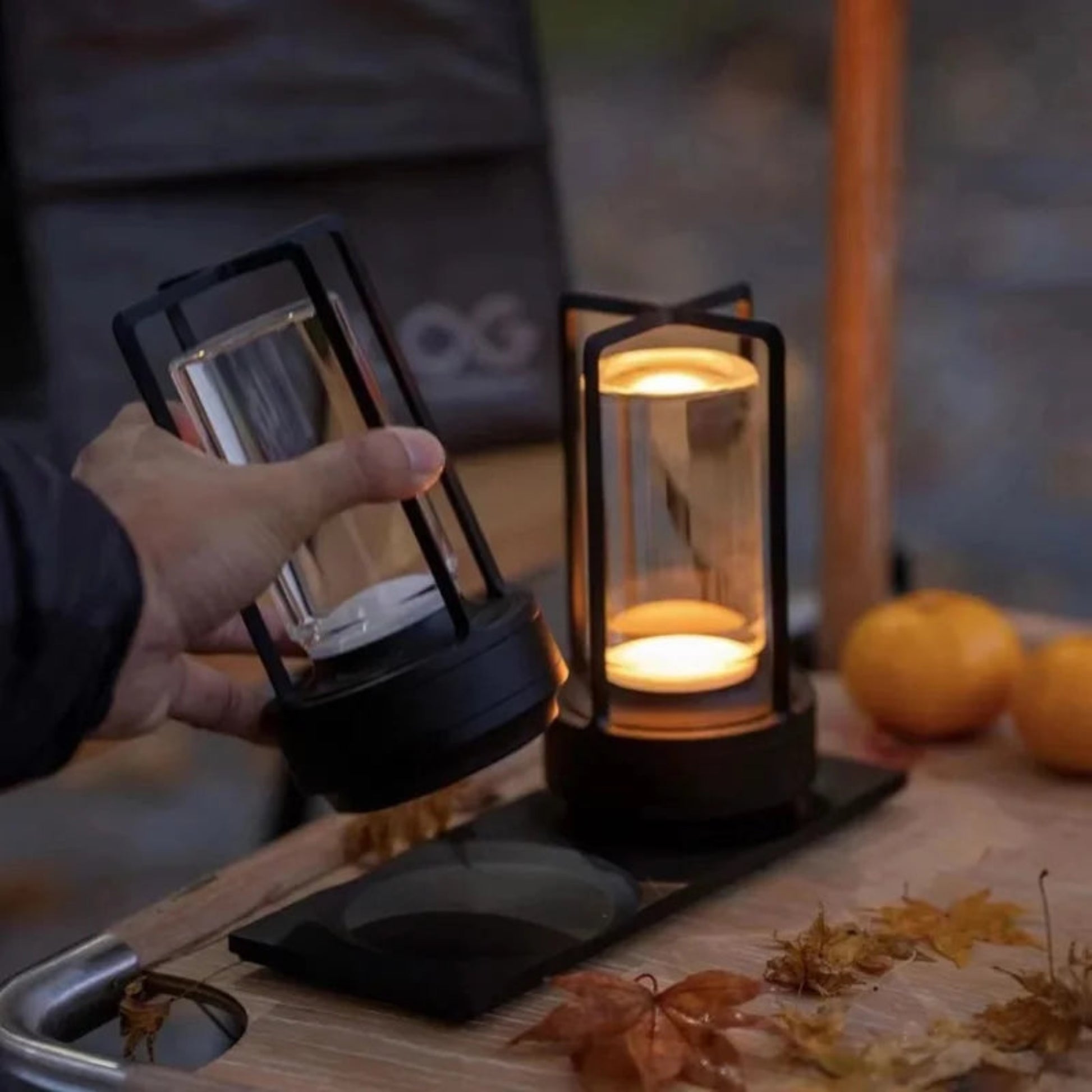 Hand holding a black lantern with another lantern on a table with pumpkins and leaves.