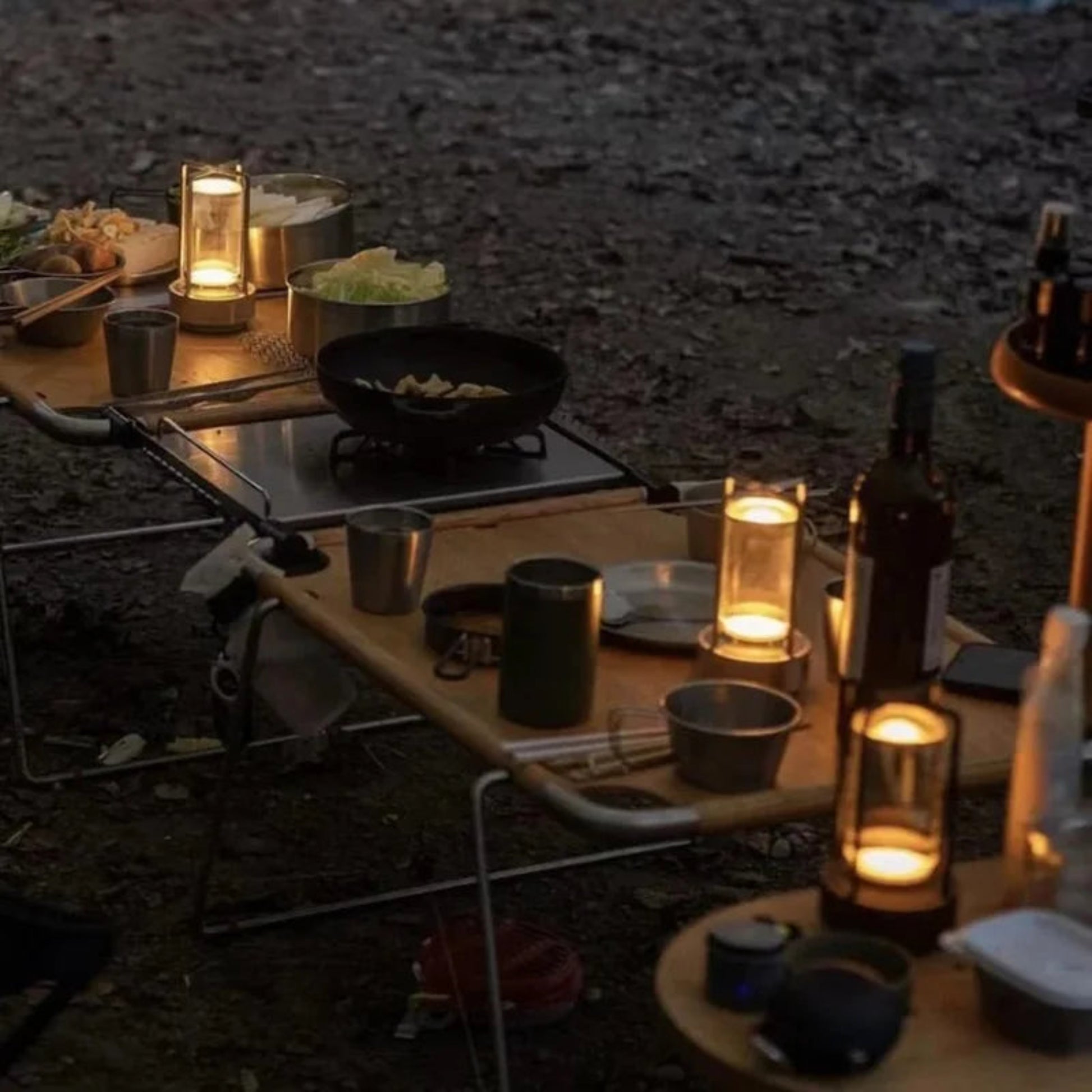 Camping setup with candles, cooking equipment, and food on a wooden table in the dark.