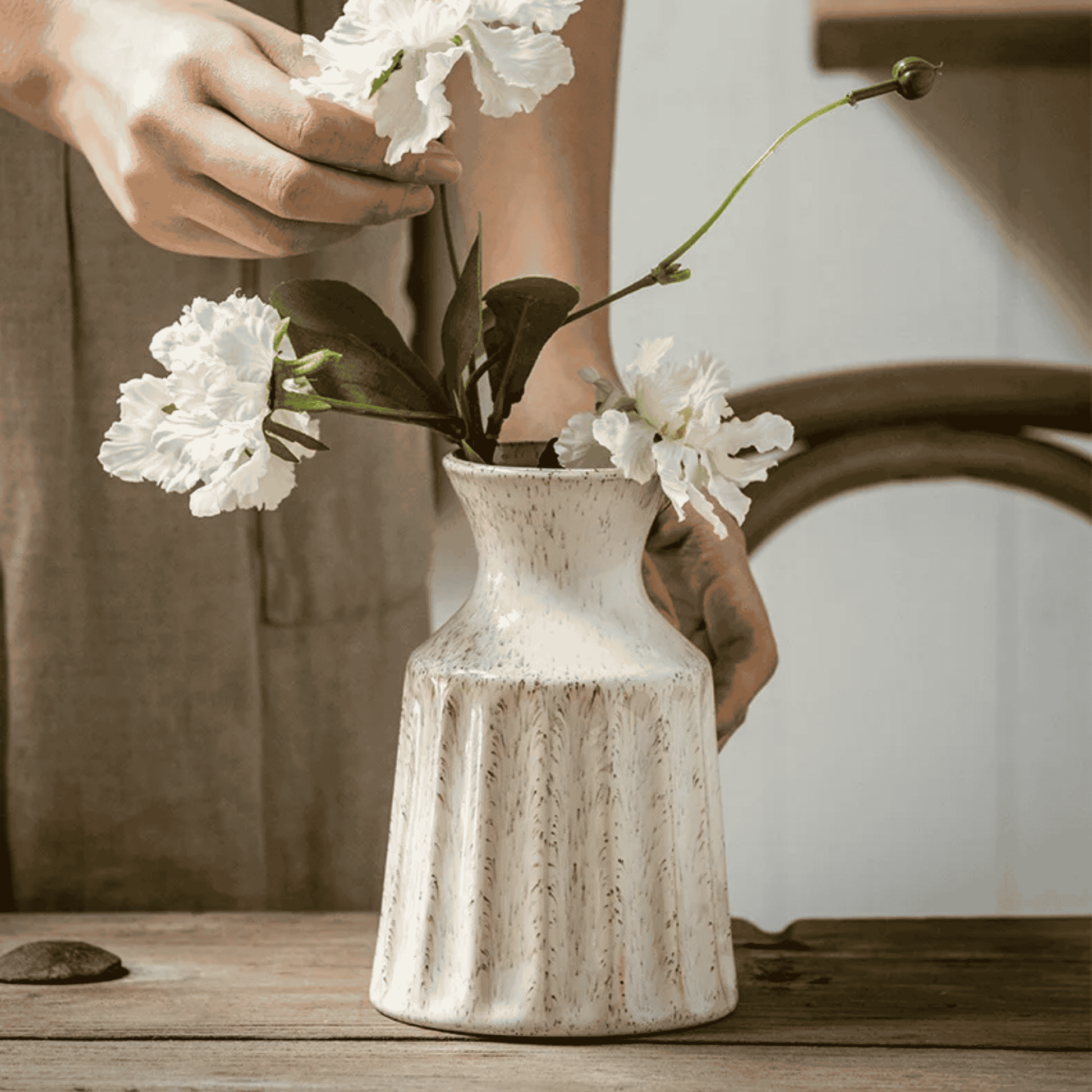 Person arranging white flowers into a rustic ceramic vase on a wooden surface.