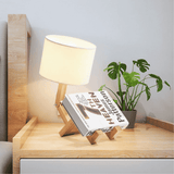 Wooden lamp with a book stand holding an open book on a wooden surface with a plant in the background.