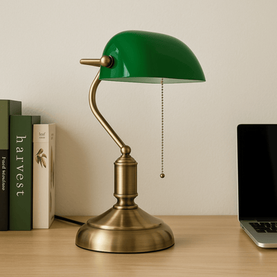 Green and brass desk lamp on a wooden surface with books and a laptop in the background.