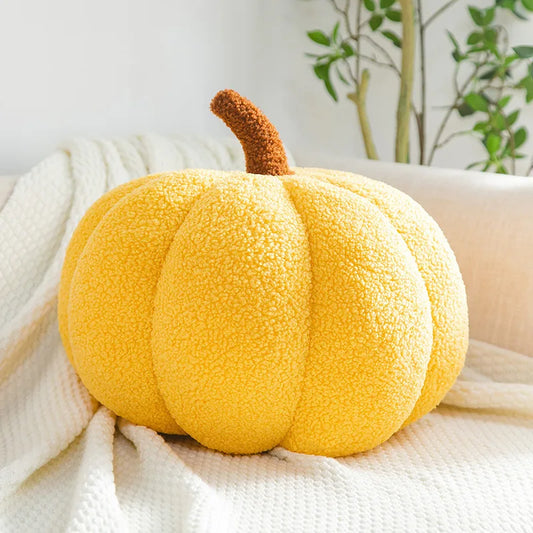 Yellow pumpkin-shaped pillow on a white blanket with a plant in the background