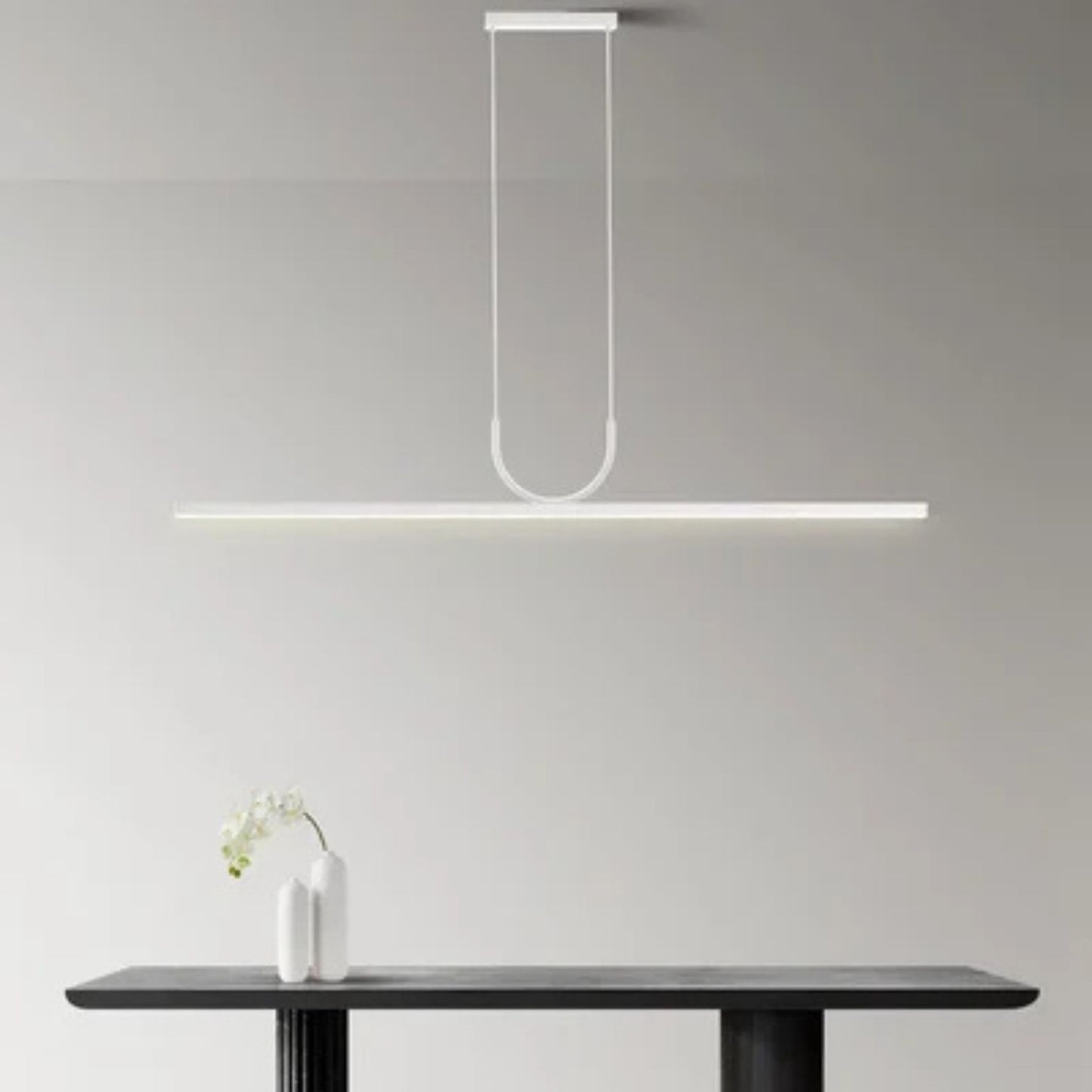A sleek, modern pendant light hangs over a dark table with a white vase and orchid, creating a minimalist, elegant atmosphere.