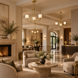 A luxurious, high-end interior lounge area featuring plush beige armchairs, a round stone coffee table with a floral centerpiece, elegant brass chandeliers, a fireplace on the left, and a reception desk in the background.