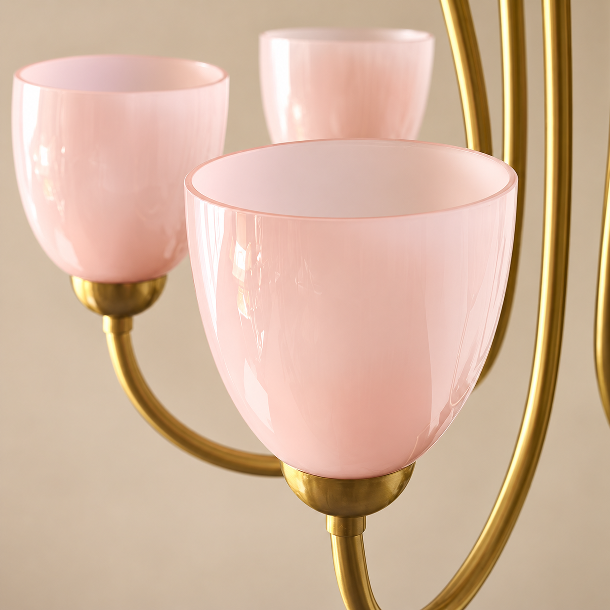 A close-up of a three-light chandelier with curved gold-finished metal arms and translucent blush pink glass shades, set against a warm beige background.