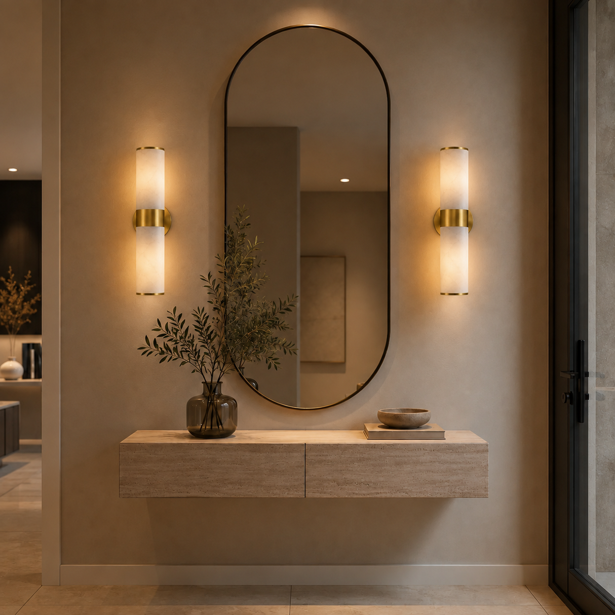 An elegant oval wall mirror with a thin black frame hangs on a beige wall above a light wood console table. Flanking the mirror are two modern cylindrical wall sconces with warm glowing glass and gold accents.