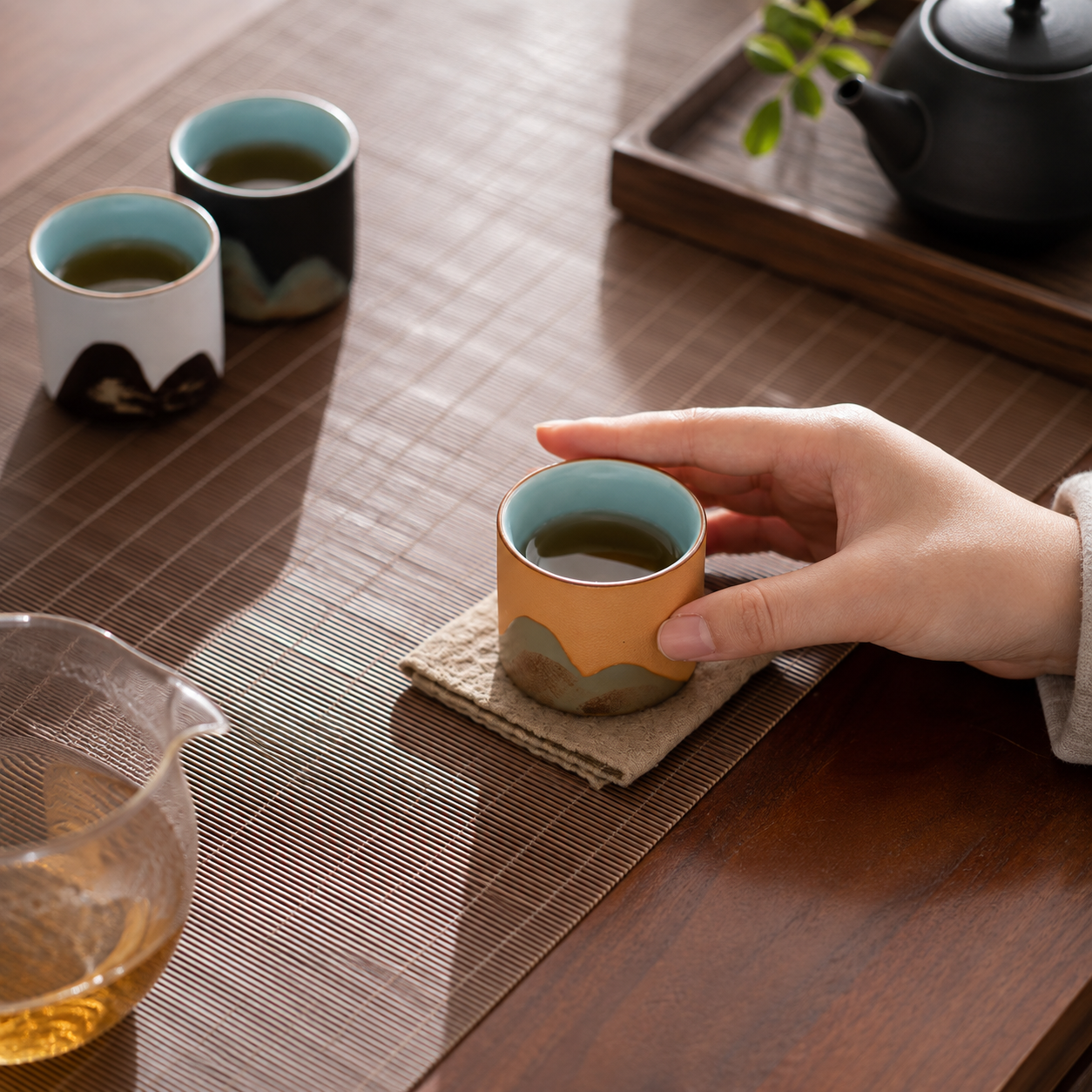 A hand holds a small ceramic tea cup with a warm orange-brown exterior and a light blue interior, placed on a woven coaster on a wooden table. Two more cups are visible in the background, along with a glass teapot and a black teapot on a tray.