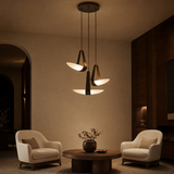 A modern three-light pendant chandelier with dark metal cone-shaped shades and glowing white oval diffusers, hanging in a warm, dimly lit living room with two cream armchairs and a round wooden coffee table.