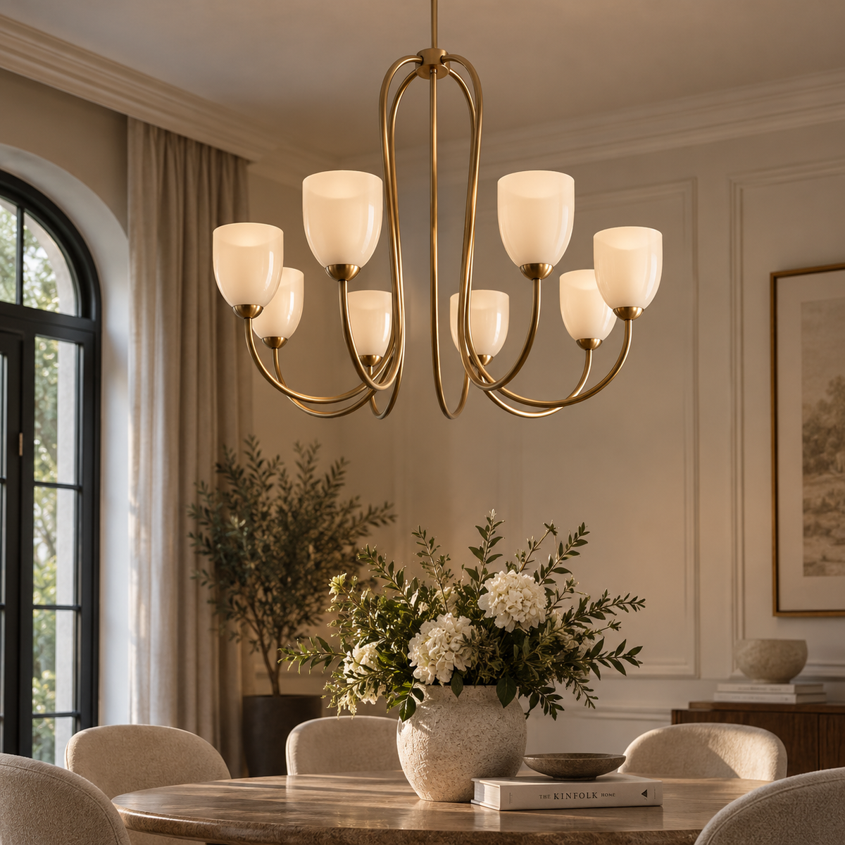 A modern multi-light chandelier with a brushed brass frame and frosted white glass shades hangs over a dining table in a styled dining room with beige walls, plants, and neutral chairs.