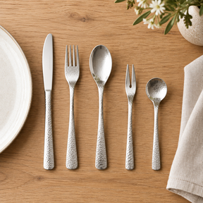 Hammered silver cutlery – 5pcs – lifestyle All selected utensils fully visible laid beside ceramic dinner plate on elegant oak dining table, linen napkin, natural daylight.